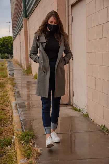 A woman wearing a black turtleneck sweater, dark wash jeans, and white sneakers, walking down a city street. She is carrying a neutral-colored tote bag over her shoulder. The scene is casual and stylish, showcasing the versatility of capsule wardrobe pieces in everyday life.