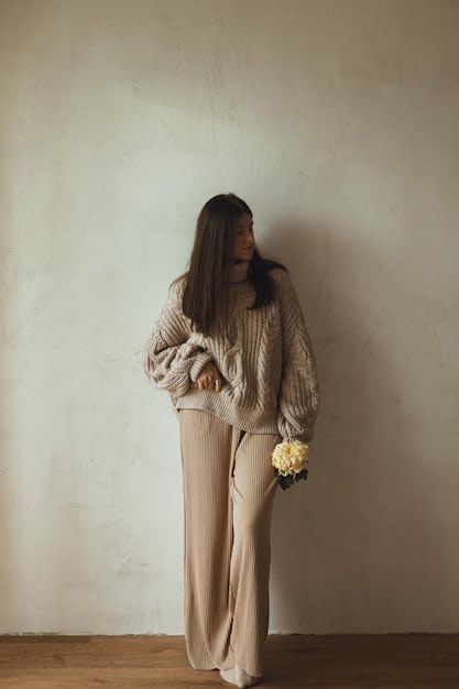 A woman wearing a monochromatic neutral outfit consisting of a gray turtleneck sweater, beige wide-leg pants, and a cream-colored coat. The look is accessorized with a brown leather belt and delicate gold jewelry.