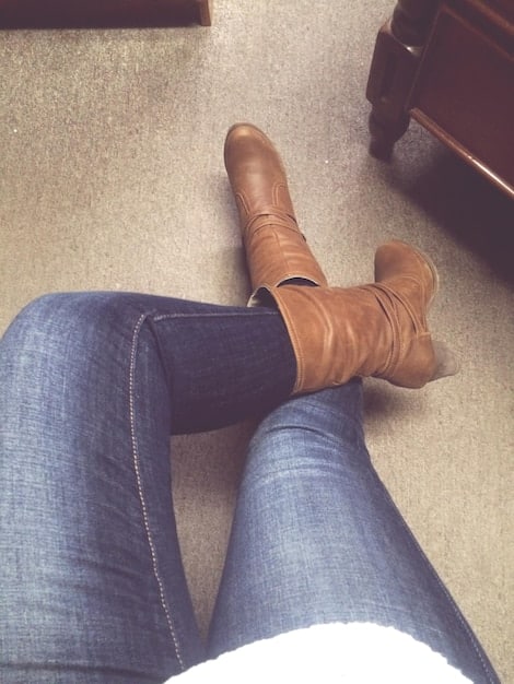 A close-up shot of a woman's feet wearing dark-wash bootcut jeans and brown leather heeled boots. The hem of the jeans slightly covers the top of the boots, showcasing a seamless and stylish transition.