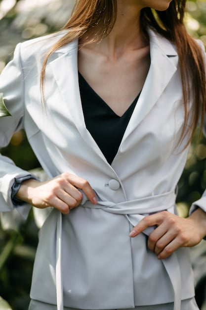 A close-up of a woman's work outfit featuring a grey blazer, a crisp white button-down shirt, dark tailored trousers, and classic black leather loafers. The outfit is accessorized with a simple silver wristwatch.