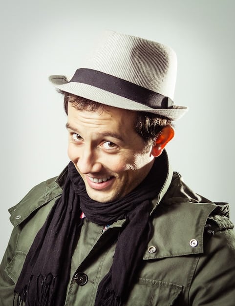 A man with a round face wearing a fedora hat. He is smiling and standing in front of a blurred background. The hat adds angles to his face.
