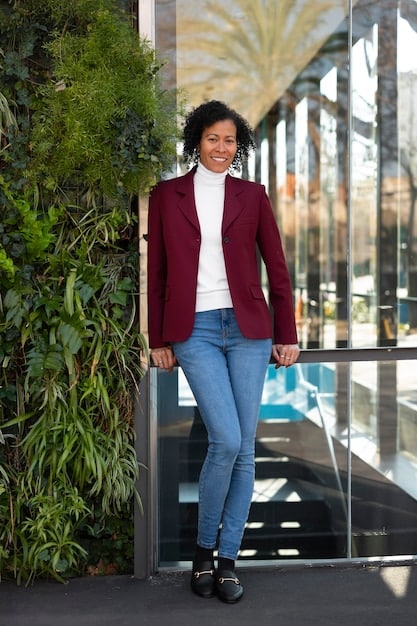 A person wearing dark-wash denim jeans paired with a white silk blouse, a black blazer, and pointed black heels, accessorized with gold hoop earrings and a delicate necklace. The backdrop is a modern office space.