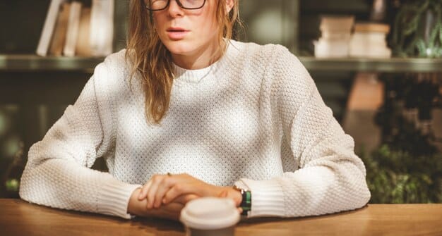 A close-up shot of a woman's hands adjusting the collar of a white button-down shirt, paired with a chunky knit sweater and gold jewelry. The background is a blurred coffee shop setting.