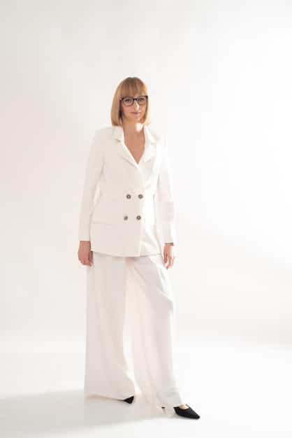 A woman wearing a head-to-toe white outfit, consisting of a tailored blazer, wide-leg trousers, and a simple t-shirt. She accessorizes with minimalist gold jewelry and white sneakers, standing against a clean, white wall.