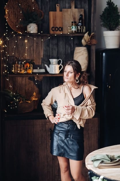 A woman wearing a tan knitted vest over a white long-sleeve shirt, paired with dark-wash jeans and brown ankle boots. The background is a cozy coffee shop with warm lighting.