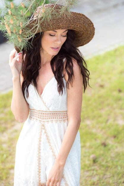 A smiling woman wearing a knee-length sundress with a light floral print, accessorized with sandals and a straw hat, attending a casual outdoor wedding.