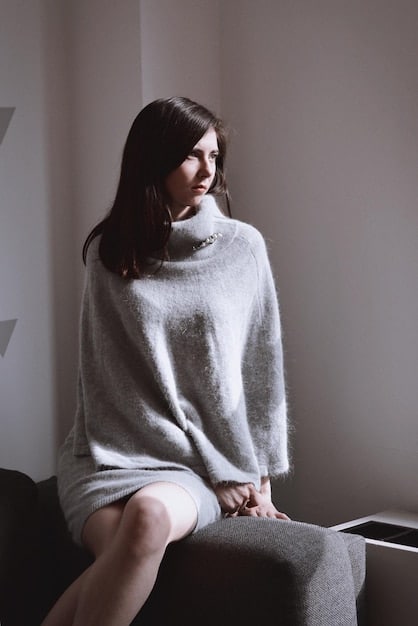 A stylish woman wearing a monochromatic gray outfit, consisting of a soft turtleneck sweater and tailored wide-leg trousers. She accessorizes with a silver statement necklace, a structured gray handbag, and white sneakers. The backdrop features an urban setting with architectural details, adding a modern, chic vibe to the overall look.