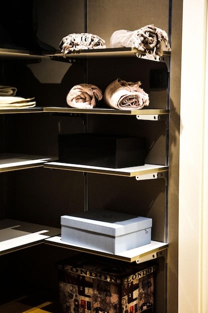 A dark closet with several shelves dedicated to perfume storage. The perfumes are arranged neatly, with some in their original boxes or opaque containers. A small dehumidifier is visible on the bottom shelf.