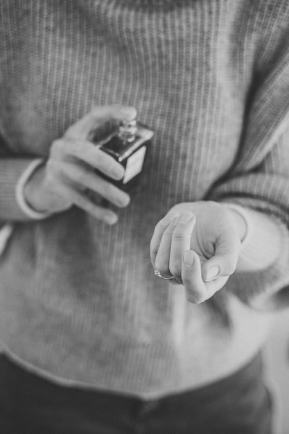 A close-up shot of a hand holding a sleek, modern-designed unisex perfume bottle with minimalist packaging, with a blurred background of various perfume ingredients like flowers, spices, and wood chips.